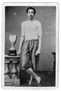 A black and white image of Arthur Wharton standing with his legs crossed next to a trophy